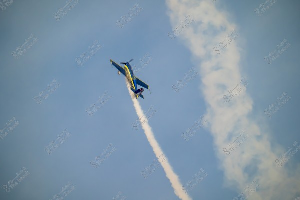 A blue, yellow, and red plane flying in the sky at an upward tilt, leaving a trail of white smoke. The sky is clear and blue with a winding smoke pattern trailing behind the plane.