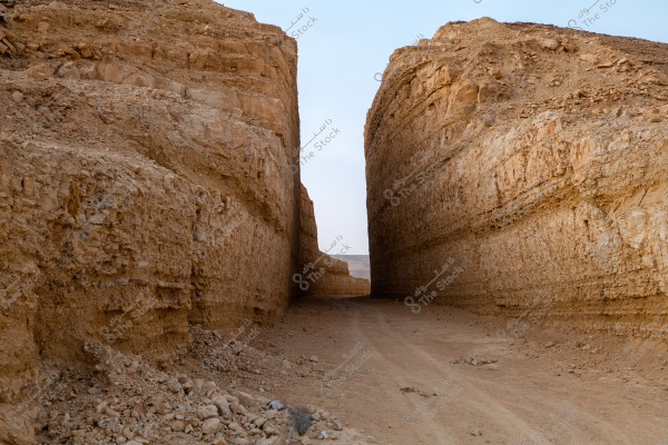 The image depicts a narrow pathway passing through a natural gorge or valley between towering, earth-brown rocky walls. The scene conveys a sense of seclusion and tranquility, with the blue sky softly illuminating the landscape. The natural details in the rocks reveal erosion and natural processes that have shaped this pathway over time.
