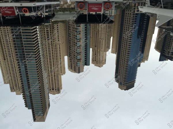 An image of a cluster of skyscrapers and modern buildings situated by a waterfront, shown upside down. The buildings have a contemporary and glassy design with multiple floors and glass balconies. At the top, there is a boat or small platform with a sign saying \"Exclusive Yachts\". The sky is cloudy and lacks any other distinct details.