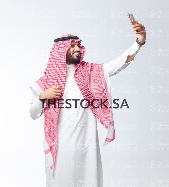 بورتريه رجل سعودي على خلفية بيضاء يقوم بالتقاط صور سيلفي وهو مبتسم ، صور تذكارية ، توثيق لحظة سعيدة