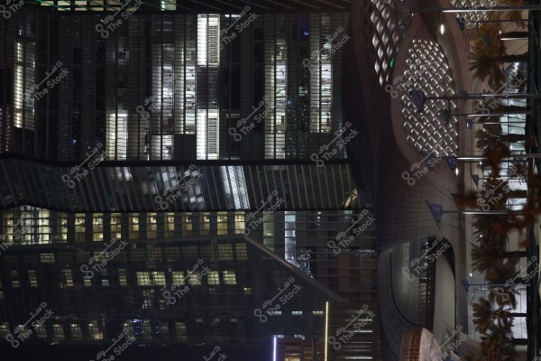 A nighttime image of a modern building with stunning architectural design and internal lighting, showcasing a large glass facade in detail. The bold architecture features curved elements with illuminated lattice openings. The building is surrounded by palm trees and flags waving in the foreground.