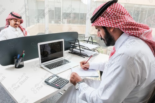 رجل سعودي يرتدي الزي السعودي التقليدي يعمل في مكتب بواجهة زجاجية ويستخدم قلم رصاص لتدوين الملاحظات