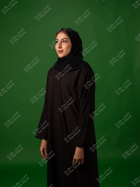 A woman wearing a black abaya and hijab stands in front of a green background.