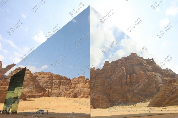 The image shows a large building with reflective glass facades, mirroring the surrounding natural scene. The building reflects rock formations and sandy mountains in the desert under a clear blue sky with scattered clouds. There are small figures of people and vehicles near the building indicating the massive scale of this structure.