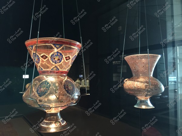 Decorated glass mosque lamps suspended by thin wires in a museum. The lamps are adorned with traditional patterns in blue, red, and gold colors. In the background, exhibit details can be seen behind a transparent glass.