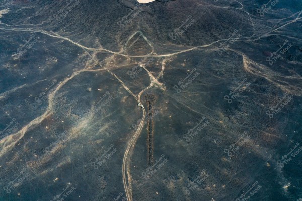 Aerial view of a desert area with rugged terrain forming random patterns. The image shows a network of dirt roads creating an irregular intersection. The ground is dark and variegated with light spots. There is no presence of dense vegetation or significant urban features near the site.