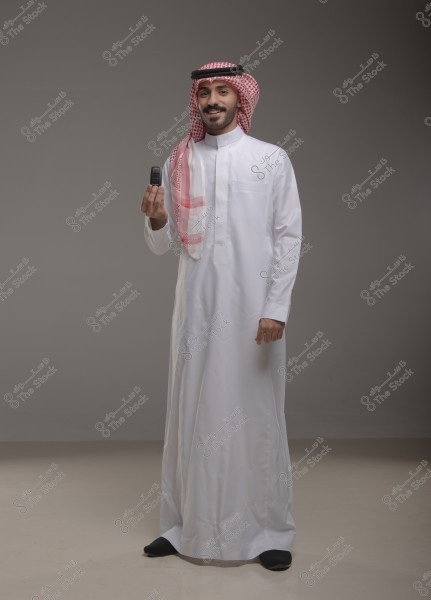 A portrait of a man dressed in traditional Saudi attire, consisting of a white thobe and a red checkered headscarf (shemagh) on his head, standing in a studio with a simple grey background. The man is holding a smart car key and has a smiling expression.