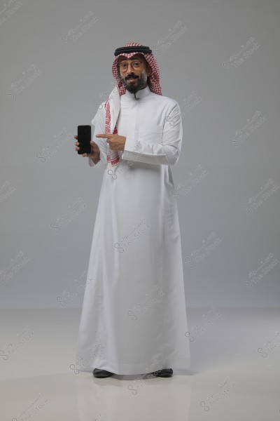 صورة لرجل يرتدي ثوبًا أبيض وغترة حمراء وبيضاء وعقال أسود. يبدو الرجل واقفًا في وضعية مستقيمة, مشيرًا بأصبعه إلى هاتف محمول يحمله بيده الأخرى. يبدو أن الصورة التقطت في استوديو بإضاءة جيدة وخلفية رمادية بسيطة.