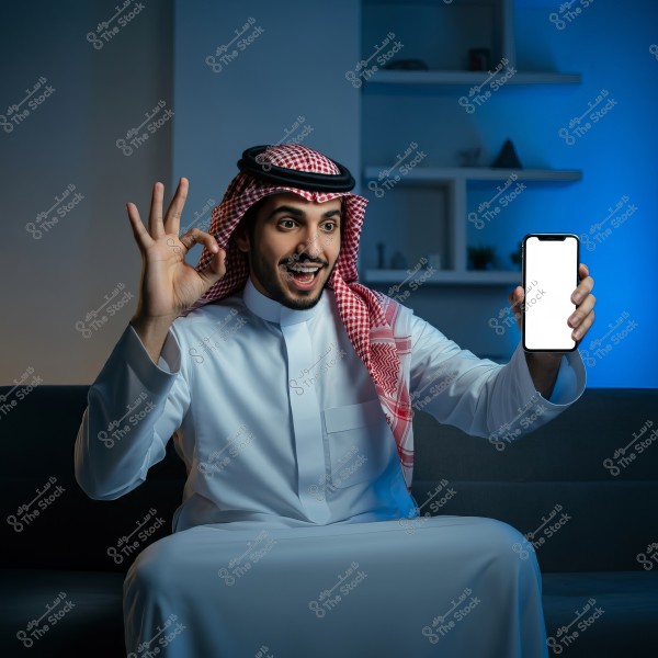 An image of a man wearing traditional Saudi attire, including a white thobe and red-and-white checkered shemagh, sitting on a couch with a smile. He is holding a smartphone with a blank white screen in one hand and showing an \"OK\" sign with the other hand. Behind him are shelves with some household items, and the background is lit with blue hues.