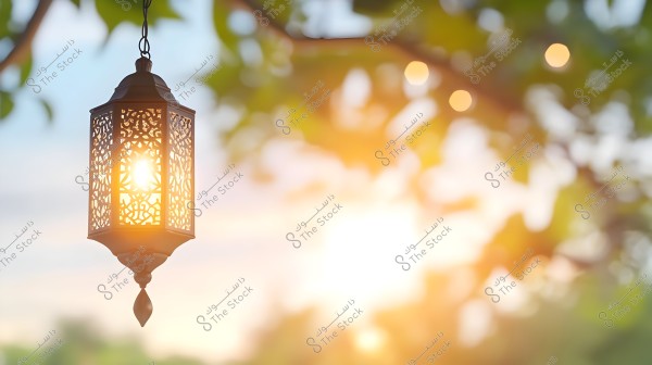 A decorative lantern in a traditional Arabic style, hanging and emitting a warm light amid a blurred background of green trees. The design features intricate geometric patterns that highlight its classic beauty. The setting appears to be in a garden or outdoor area during sunset, creating a tranquil and enchanting atmosphere.