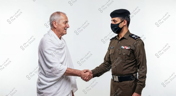 The image shows two people shaking hands against a white background. The person on the left is wearing a white Ihram, typically worn for the Hajj pilgrimage. The person on the right is dressed in a dark brown military uniform with a matching cap and a black face mask. The scene connects security and spirituality, possibly indicating a setting in Saudi Arabia.