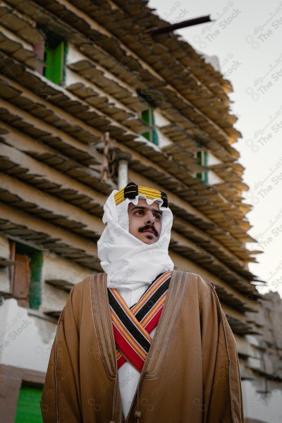 A snapshot of a Saudi man wearing a popular heritage costume on the day of the foundation stands in front of an clay building, heritage uniform, foundation day, old archaeological buildings