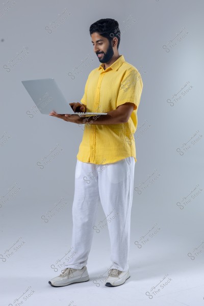 A man stands in front of a gray background holding a laptop and appears happy. He is wearing a yellow shirt, white pants, and white sneakers. The image shows him in a side profile while working on the device.