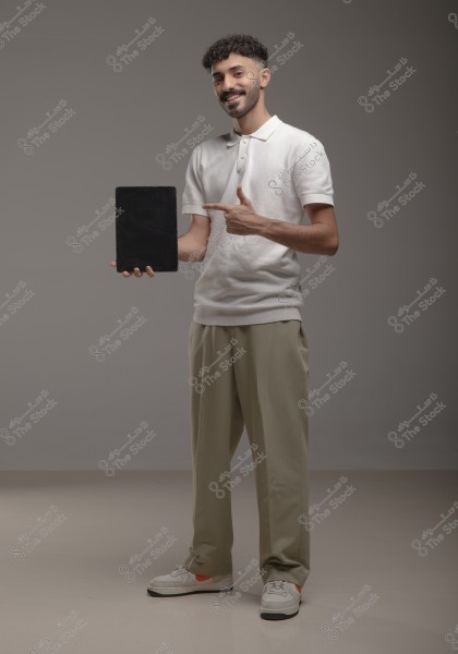 A portrait of a man standing in a minimalist studio, smiling at the camera. He is wearing a white polo shirt, beige trousers, and white sneakers. He holds a tablet in one hand and points to it with the other. The grey background highlights the subject of the photo.