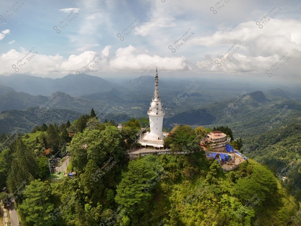 An aerial view of a tall white tower situated on top of a hill covered with green trees. A stunning vista of mountains extends in the background under a partly cloudy sky. Some small buildings are nearby the tower, with blue tarps covering parts of the surrounding areas.