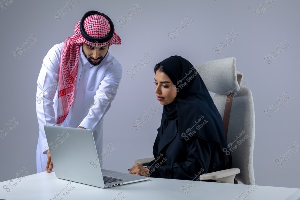 An image of a man and woman in an office setting. The man is wearing a white thobe and a red and white checked headscarf, typical of Saudi attire, and is pointing towards a laptop screen. The woman is seated in an office chair, wearing a black abaya and hijab, attentively looking at the laptop. The background is light gray, adding a professional atmosphere.