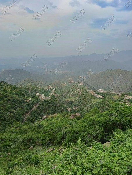 A natural landscape of green mountains in a mountainous region, featuring scattered houses among dense trees. The horizon has a light mist, and the sky is partially covered with clouds.