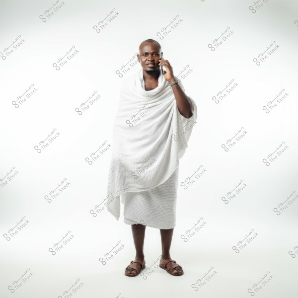 A man stands in a portrait pose, wearing traditional white Ihram clothing. He is holding a mobile phone to his ear, appearing to be in conversation. The background is plain white, and he is wearing brown sandals.