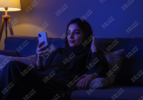 A portrait of a woman sitting on a sofa in a dimly lit room with a floor lamp in the background, wearing a black abaya and hijab, using a smartphone. The lighting creates a calm and cozy atmosphere, and the woman appears relaxed and engaged.