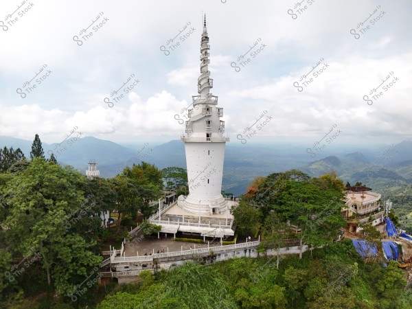 The image shows a tall white tower with a spiral design at the top, surrounded by dense green trees. The scene extends to mist-covered mountains in the distance with white clouds in the sky. There is a white railing around the base of the tower, with a building beside it.