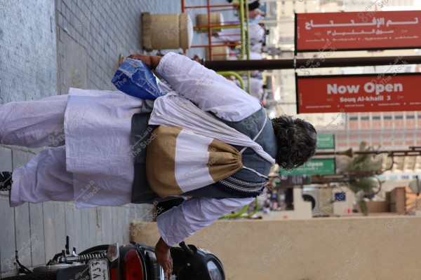 A man walks along a sidewalk in a city, wearing a white robe and a gray vest, carrying a cloth bag on his back and a blue plastic bag in his hand. A sign next to him reads \"Now Open\" and \"Souq Al-khalil 3.\" The area is bustling with passersby and adorned with signs in Arabic and English.