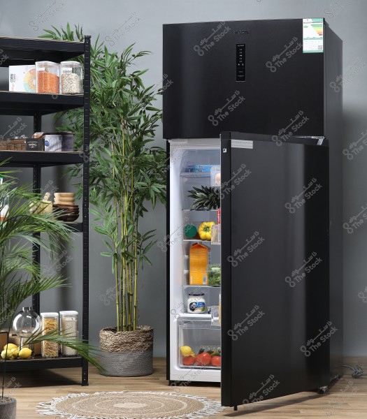 The image depicts a modern and stylish kitchen. A black refrigerator is open, displaying various items like eggs, milk, vegetables, and fruits. Next to the fridge, tall green plants are in an attractive pot. To the left, there is a black shelving unit holding storage containers and food items, including pasta and grains. The floor is wooden, and there is a small circular rug in the center.