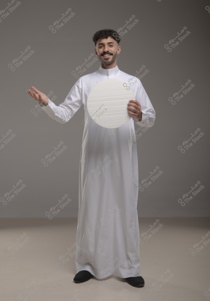 صورة لرجل يقف في استوديو مع خلفية رمادية. يرتدي الرجل ثوباً أبيض طويلاً, ويبدو أنه من الطراز العربي التقليدي, مما يشير إلى أنه ربما من السعودية أو إحدى دول الخليج. يمسك الرجل بيد واحدة دائرة بيضاء ويبدو مبتسماً.