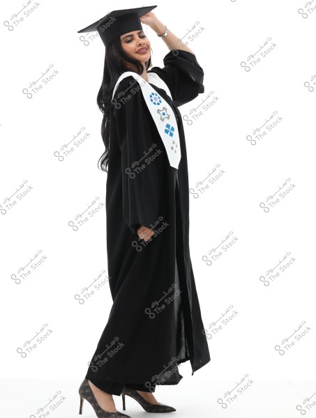 Image of a smiling woman wearing a black graduation gown and mortarboard. The gown is adorned with a white stole featuring geometric symbols in various colors like blue and gray. The woman is wearing high-heeled shoes and is standing sideways, holding the edge of her cap with her hand.