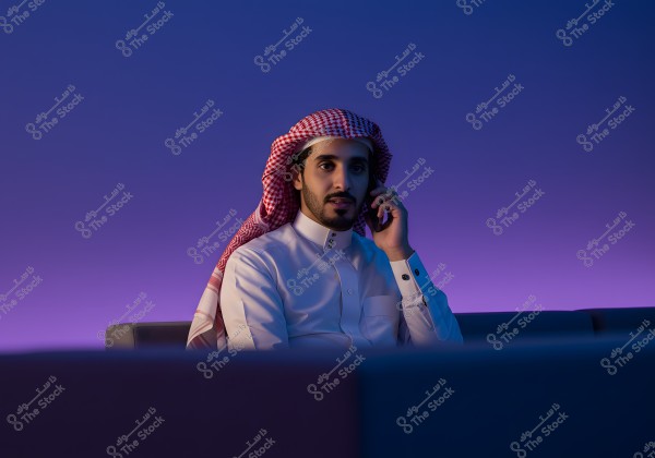 Image of a man wearing traditional Saudi attire, including a white thobe and a red and white checkered ghutra, sitting on a sofa and talking on a mobile phone. The background is illuminated with blue and purple lighting.