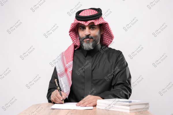 A man wearing traditional Saudi thobe and ghutra sits at a table writing with a pen, and there are books in front of him.