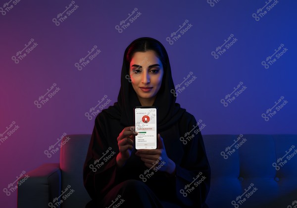 An image of a woman sitting on a sofa, wearing a black abaya and hijab, holding a smartphone with a bright screen displaying an app. The background is shaded in blue and purple hues.