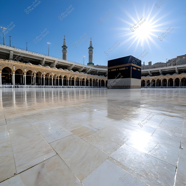The image shows the Kaaba in Mecca, Saudi Arabia, under a clear blue sky with a bright sun. Surrounding the Kaaba are clean, shiny tiles, and the distinct architecture of the Grand Mosque with its arched columns is visible in the background.