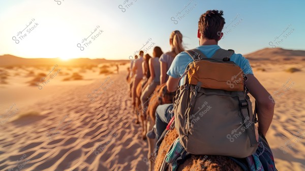 A group of people riding camels in a single file during sunset in an open desert. The golden rays of the sun are visible on the horizon, with a clear sky. The people are dressed casually, and the desert sand landscape is visible around them.