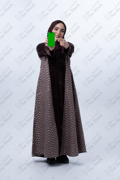 A young Saudi woman wearing an abaya and hijab, holding a green-screen phone