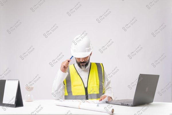 The image shows an engineer or construction worker seated at a desk. He is wearing a white safety helmet and a yellow reflective safety vest over traditional white thobe attire. He is holding a pen and reviewing some blueprints in front of him. On the desk, there is a laptop, a desk calendar, and an hourglass.