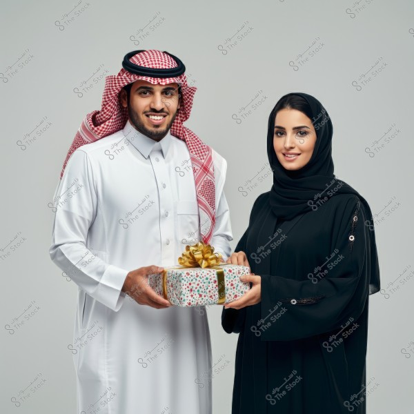 Image of a man and woman standing together, the man wearing traditional Saudi attire with a thobe, ghutra, and agal, while the woman wears a black abaya and hijab. They are holding a gift wrapped in colorful paper and tied with a golden ribbon. Their expressions are happy, and the background is a neutral gray color.
