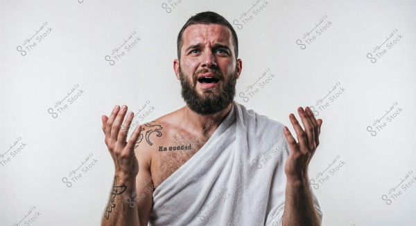 An image of a man with a short beard, wearing a white cloth draped over his shoulder, displaying an expression of surprise or prayer. The man has tattoos on his arm and chest.