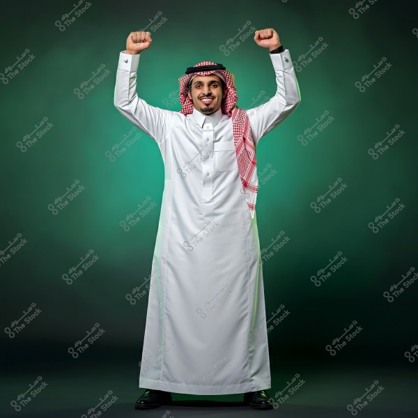 A portrait of a man standing against a green background, raising his arms proudly. The man is wearing a traditional white thobe and a red-and-white checked shemagh with an agal, indicating Saudi attire.