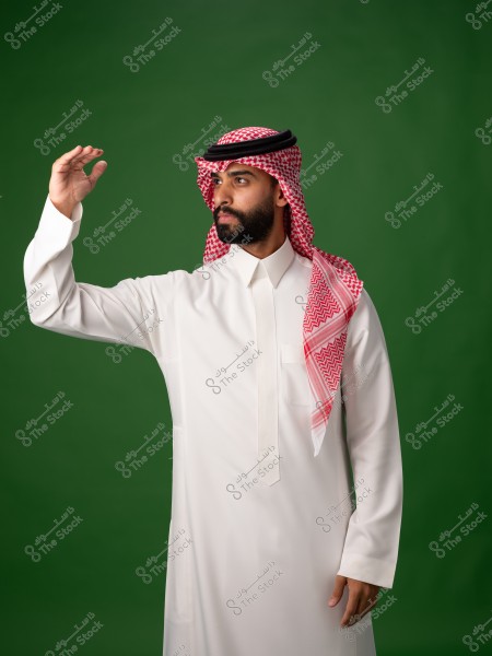 صورة لرجل يرتدي ثوباً أبيض تقليدياً وعقال وشماغ أحمر وأبيض على رأسه, يقف أمام خلفية خضراء. يبدو وكأنه ينظر بعيداً ويرفع يده كما لو كان يشير أو يحمي عينيه من الشمس.