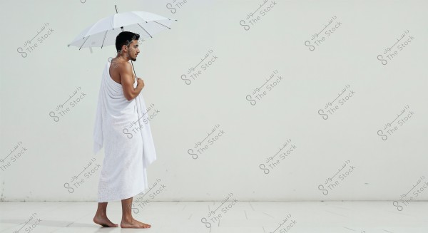 Image of a man standing barefoot on a white floor, wearing a white wrap and holding a white umbrella. The man is looking to the left side, and the background is a plain white wall.