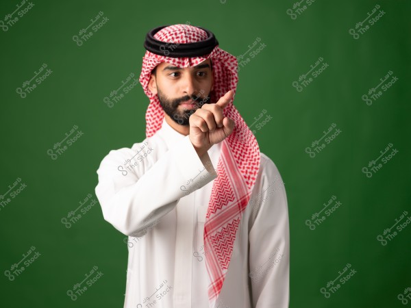 صورة لرجل يرتدي ثوبًا أبيض وغترة حمراء مع عقال أسود, يقف أمام خلفية خضراء. يظهر الرجل وهو يشير بإصبعه السبابة إلى الأمام. الزي تقليدي من منطقة السعودية.