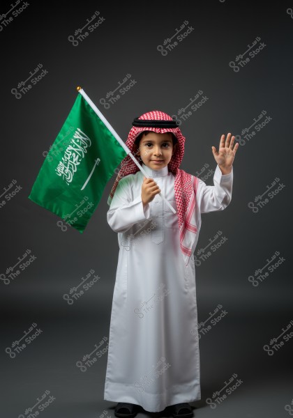 An image of a young boy wearing traditional Saudi attire, consisting of a white thobe, red checkered keffiyeh, and black agal. He is holding the Saudi Arabian flag in his right hand and raising his left hand in a greeting gesture. The background is dark, emphasizing the details of the boy and the flag.
