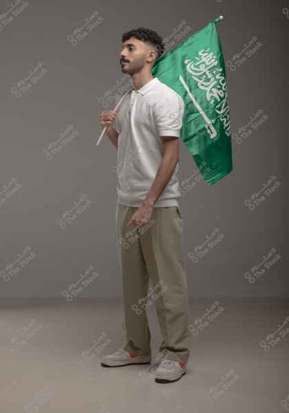 A portrait of a man holding the flag of Saudi Arabia. The man is wearing a white polo shirt and beige trousers paired with white sneakers. He is standing in a side pose against a gray background.