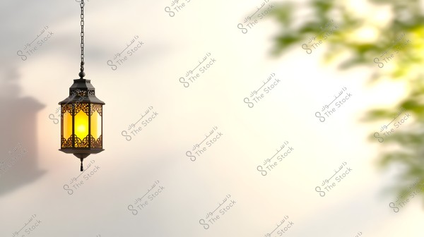 صورة لفانوس معدني تقليدي معلق بسلسلة, مضاء بضوء دافئ يشع للخارج, يشكل ظلالاً على الجدار. يظهر في خلفية الصورة تدرجات لونية هادئة وأوراق شجر خضراء على الجانب الأيمن.