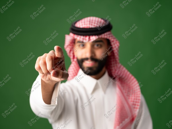A portrait of a man wearing a white thobe, a red and white checkered keffiyeh, and a black agal, standing against a green background, pointing forward.