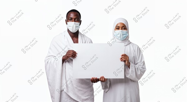 An image of a man and a woman wearing medical masks, holding an empty white board. The man is dressed in a white ihram, suggesting he may be on a pilgrimage or Umrah, while the woman is wearing a white hijab and dress, reflecting traditional Islamic attire. Both are set against a white background.