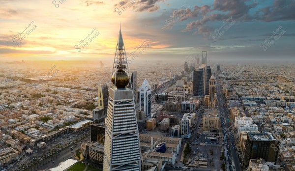 An aerial view of Riyadh city, the capital of Saudi Arabia, featuring a cluster of high-rise towers including the iconic Al Faisaliah Tower with its pyramid structure and glass orb at the top. In the background, the Kingdom Tower with its distinctive sky bridge is visible. The sky is adorned with the stunning colors of a sunset, showcasing hues of orange and blue, while the vast cityscape stretches underneath.