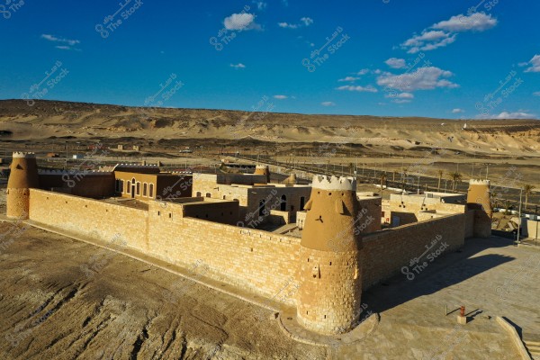 An image of a traditional fortress made of stone and adobe, featuring round towers with jagged tops. The fortress is situated in a desert area with a backdrop of sand dunes and mountains. The sky is blue with scattered clouds, providing a cultural and historical setting.
