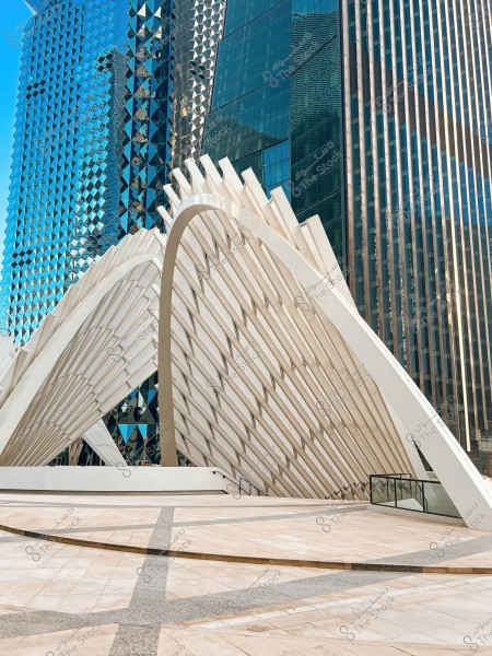 An image of a modern building with a unique architectural design featuring white curved structures and mirrored glass surfaces. In the background, there are skyscrapers with geometric designs reflected on the glass windows, giving the scene a futuristic feel. The foreground shows light-colored marble tiles with distinct pathways.