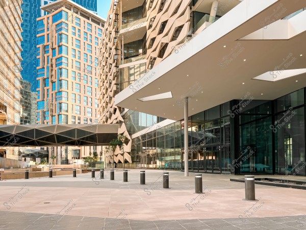 An image of an urban scene during the day, showcasing modern architecture. The buildings have glass facades and intricate geometric designs. The scene includes open public walkways and evenly distributed light poles. In the background, tall buildings with distinctive glass facades are visible under a clear blue sky.
